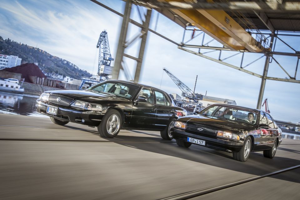 Two black sedans driving on a road near a harbor with cranes