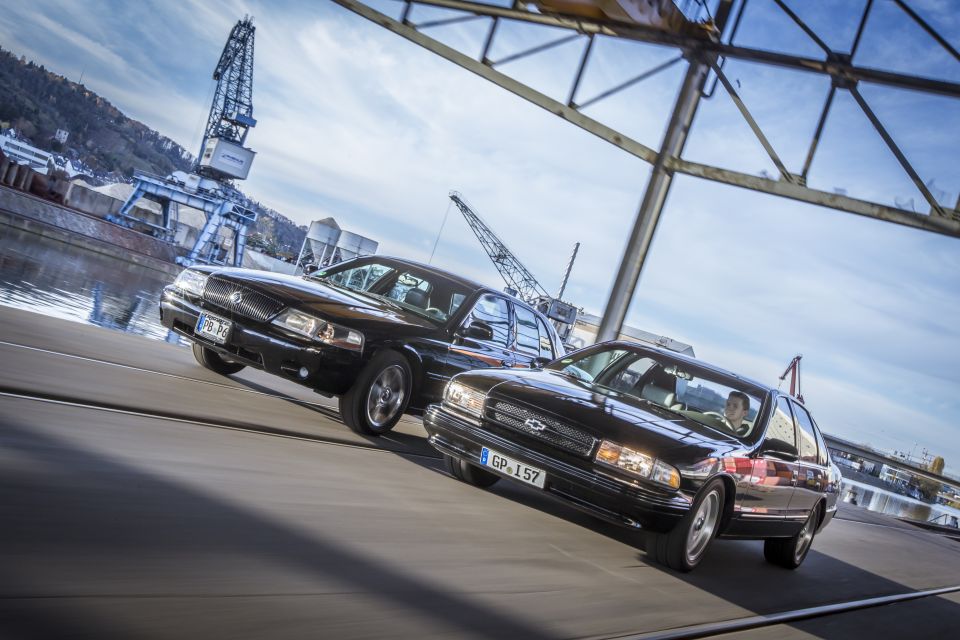 Two black sedans, one Mercury Marauder and one Chevrolet Impala, driving on a road with industrial cranes in the background