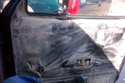1995 Ford Bronco - Inside of a door trim panel that has been burned and melted, with charred black plastic, a scorched speaker grille and torn openings where…