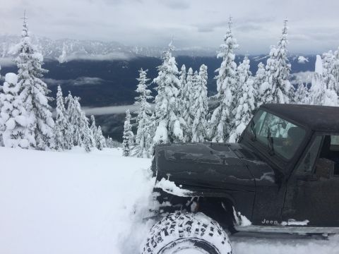 1995 Jeep Wrangler - A black Jeep navigates a snowy mountain landscape with snow-covered trees and cloudy skies.