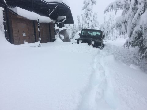 1995 Jeep Wrangler - A dark-colored Jeep Wrangler is partially buried in deep snow next to a wooden building, with snow-covered trees in the background.