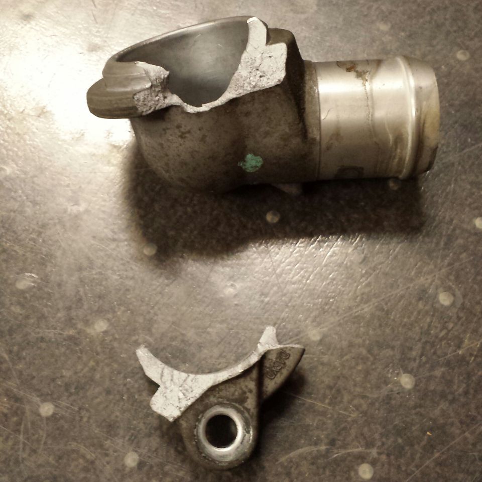 Broken metal automotive part, possibly a thermostat housing, on a textured surface.