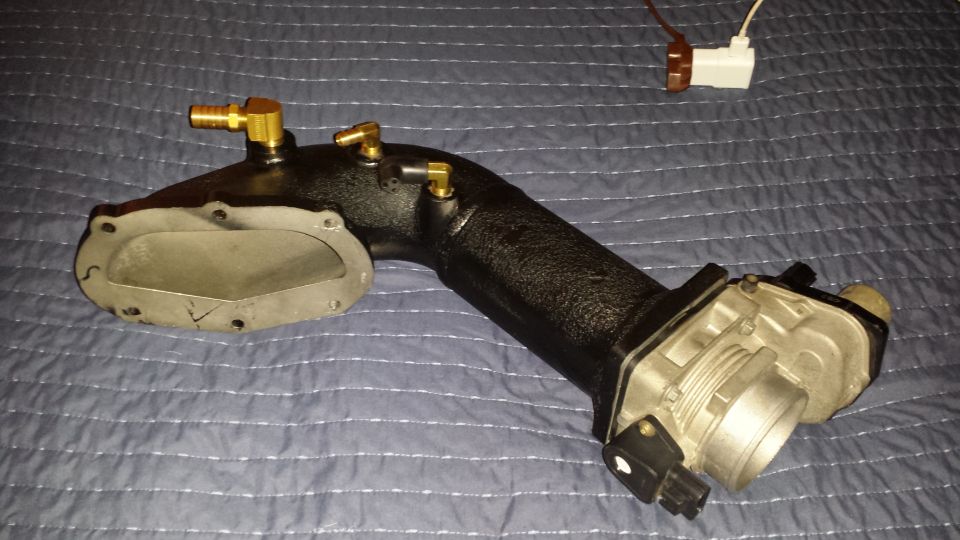 Black metal intake manifold with brass fittings and a silver throttle body, resting on a blue quilted surface