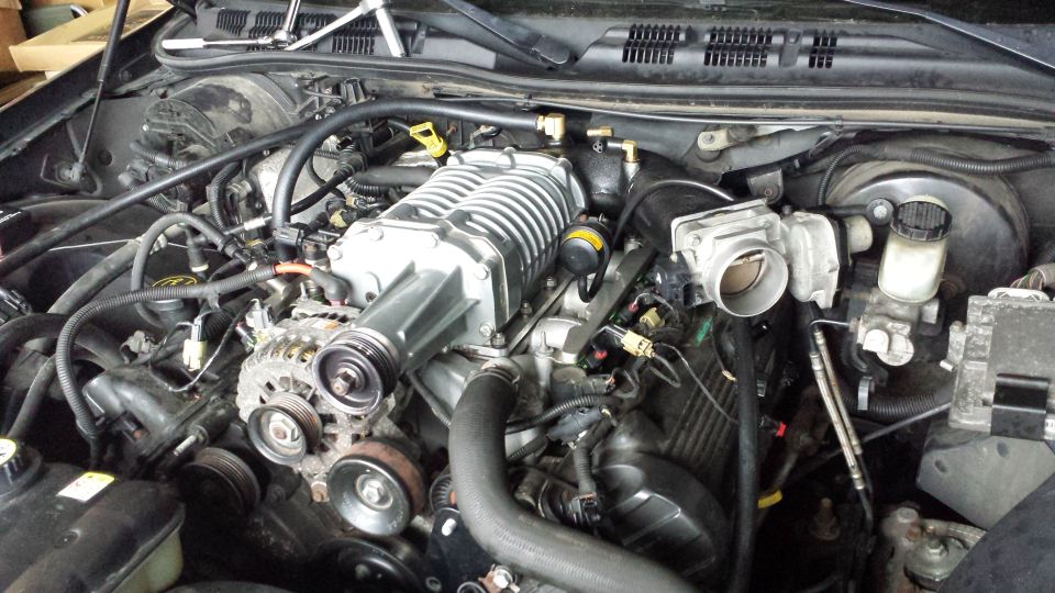 Engine bay with a prominent silver supercharger and various hoses and belts