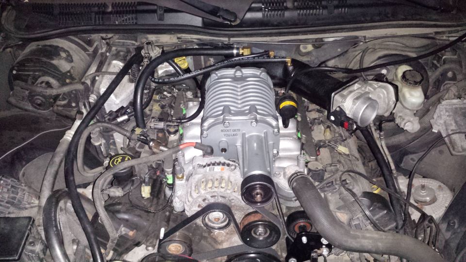 Silver supercharger installed on a car engine with visible hoses and belts.