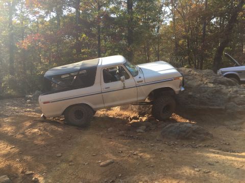 1979 Ford Bronco - A white Ford Bronco is navigating a rocky trail, with its front tires on a large boulder. The vehicle has a soft top and appears to be a co…