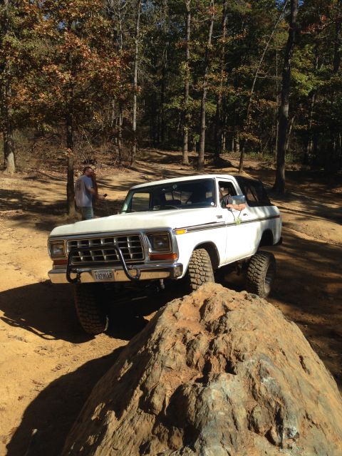 1979 Ford Bronco - A white 1979 Ford Bronco is shown off-road, navigating over a large rock with a person standing behind it. The vehicle has large tires and…