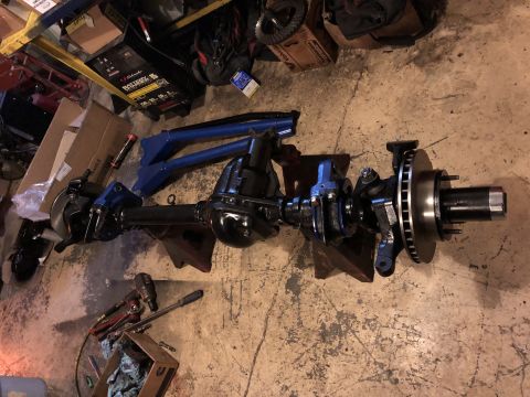 1995 Ford F-150 - A black front axle assembly with disc brakes and suspension components is displayed on jack stands on a concrete floor, with tools and part…