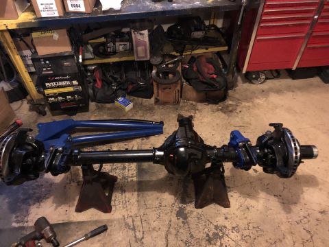 1995 Ford F-150 - A black rear axle assembly with blue suspension components is supported by two red jack stands on a concrete floor. Tools and automotive pa…