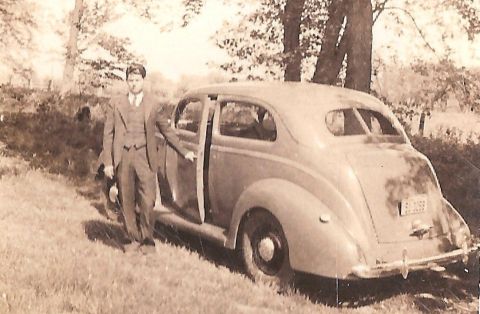 1967 Ford Custom Wagon - A man in a suit stands next to the open driver's side door of a light-colored, four-door sedan parked on a grassy slope with trees in the b…