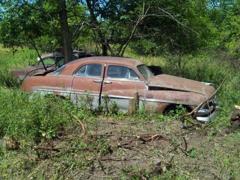 1967 Ford Custom Wagon - Two rusty, vintage sedans are parked in a grassy, overgrown area with trees. The front sedan is a dark red with a chrome bumper and hood or…