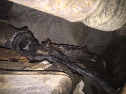 1967 Ford Custom Wagon - Close-up view of rusted linkage components attached to a vehicle's undercarriage, with a black protective boot and wiring harness visible.