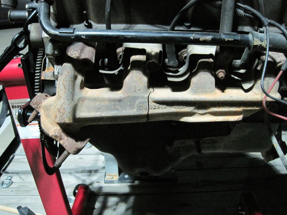 Rusty V8 engine exhaust manifold and intake on engine stand, showing casting numbers and a crack.