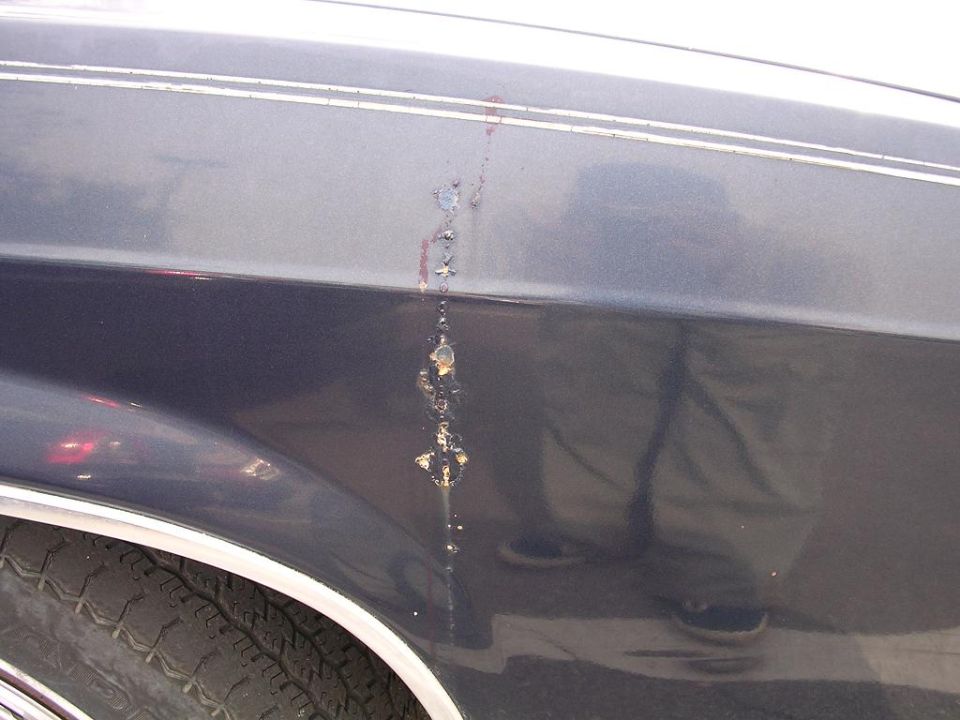 Close-up of rust spots and paint damage on dark blue fender of a sedan, above the front wheel