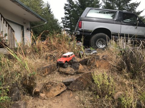 1989 Ford Bronco - RC trucks navigate a rocky terrain in front of a house and a larger vehicle.