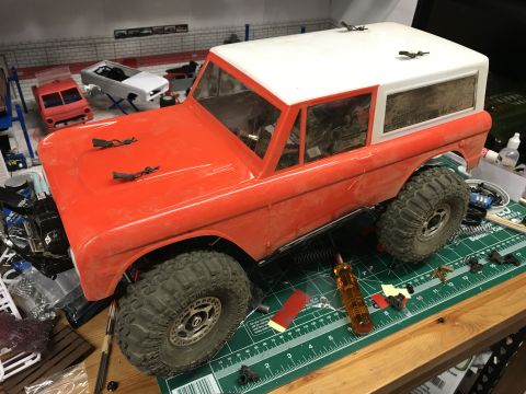 1989 Ford Bronco - An orange RC Bronco model is partially assembled on a workbench, surrounded by tools and other model car parts.