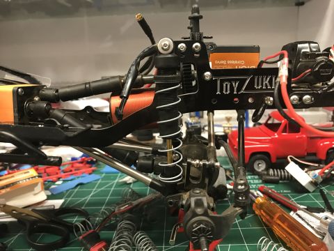 1989 Ford Bronco - Close-up of an RC car's suspension system with a red toy truck visible in the background.