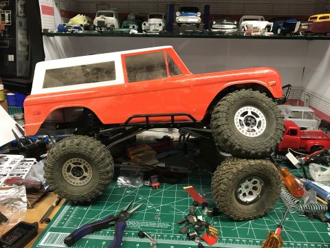 1989 Ford Bronco - An orange and white RC Bronco body sits on a chassis with large tires, surrounded by hobby tools and parts.