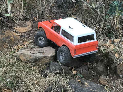 1989 Ford Bronco - An orange and white remote-controlled rock crawler navigates a rocky, outdoor terrain.