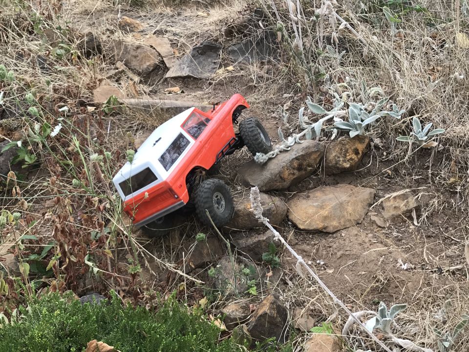 Orange and white RC truck with large tires climbing over rocks on a dry, grassy slope