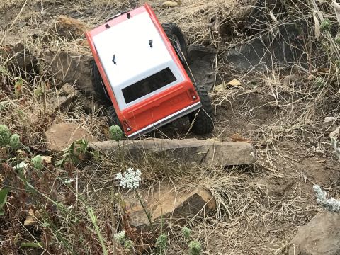 1989 Ford Bronco - An RC rock crawler truck navigates a rough, outdoor course with dry grass and rocks.