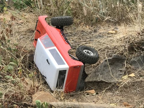 1989 Ford Bronco - An overturned red and white RC rock crawler lies on its side amidst dry vegetation and rocks.