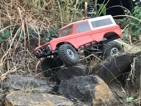 1989 Ford Bronco - An orange remote-controlled rock crawler truck navigates a rocky terrain with dry vegetation.