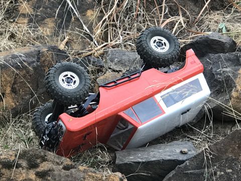 1989 Ford Bronco - An RC rock crawler, painted red and silver, is overturned on a rocky terrain, its wheels in the air.