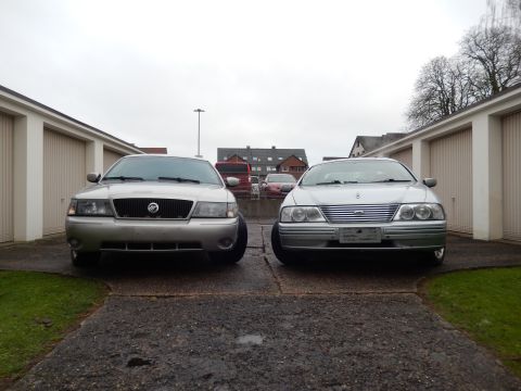1999 Ford Fairlane - Two sedans are parked in front of garages on a cloudy day.