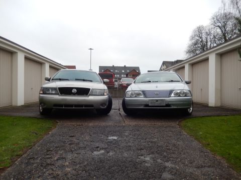 1999 Ford Fairlane - Two sedans are parked side-by-side in front of a row of garages on an overcast day.