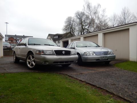 1999 Ford Fairlane - Two silver sedans, a Mercury Grand Marquis and a Ford Falcon, are parked in front of a row of garages.
