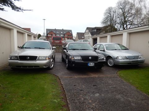 1999 Ford Fairlane - Three sedans and a wagon are parked in a driveway in front of garages.