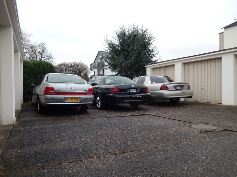 Three sedans parked in a driveway next to a house with garages. One is black, two are silver