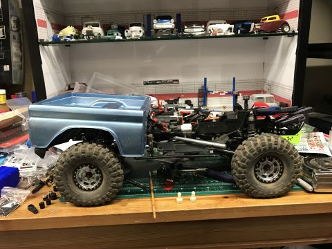 1989 Ford Bronco - An RC model truck chassis with a light blue pickup truck body is shown on a workbench, surrounded by hobby supplies.