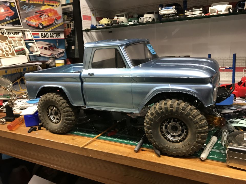 Blue model truck with large tires sits on a workbench next to tools and model kits