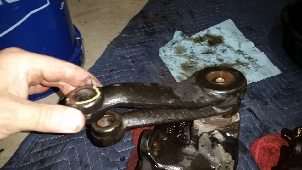 Hand holding a grimy black steering arm with bushing bores over a shop blanket during disassembly