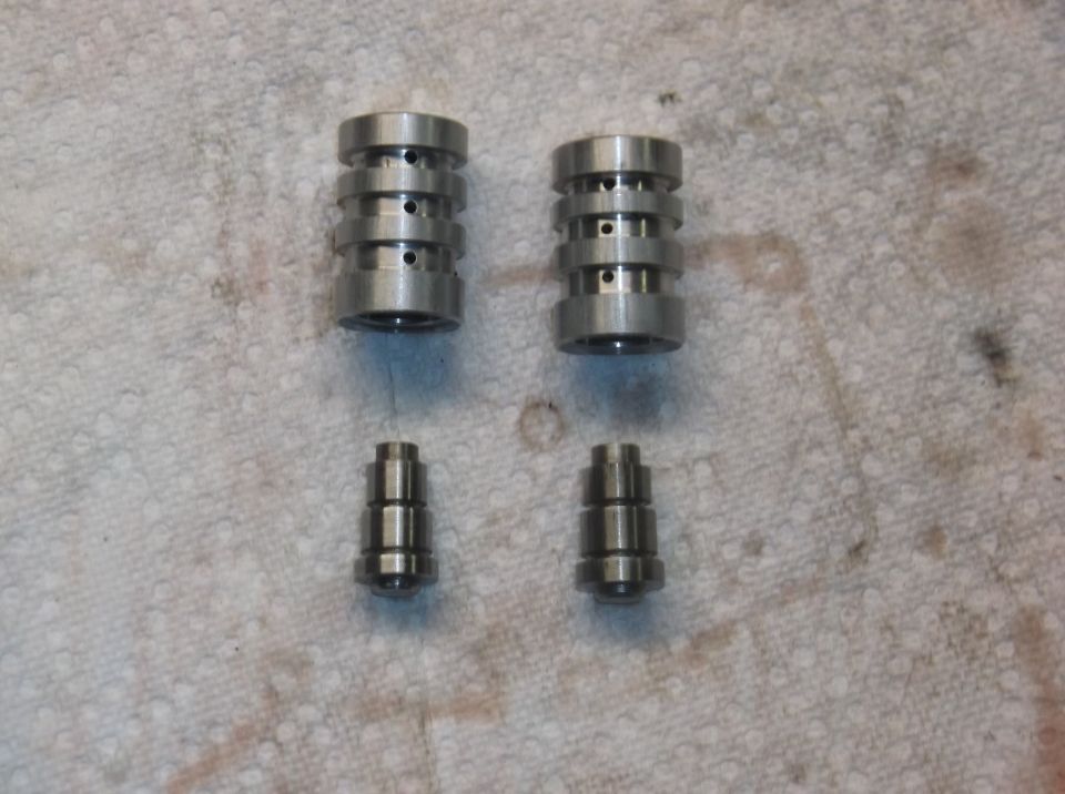 Four metal automotive engine parts, two larger rocker arms and two smaller valve lifters, laid out on a textured cloth