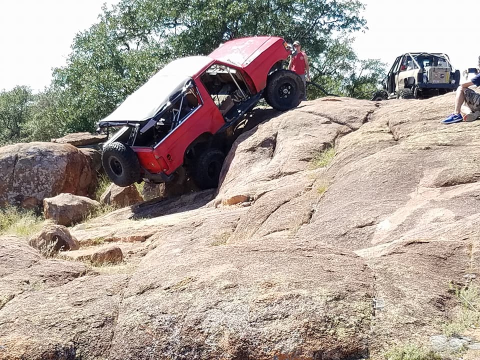 1989 Ford Bronco modified for off-roading climbing a rocky incline, another modified vehicle in background
