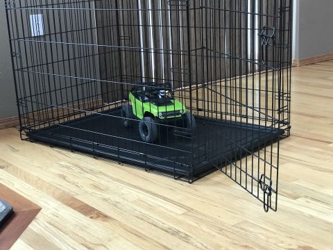 1989 Ford Bronco - A green RC rock crawler toy is inside a large black dog crate on a wooden floor.