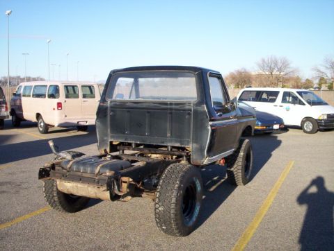 1978 Ford Bronco - Rear three-quarter view of a dark-colored, lifted early Ford Bronco with its rear body/bed removed, exposing the bare frame, fuel tank, and…