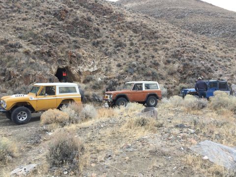 1969 Ford Bronco - Three Ford Broncos and a blue SUV are parked on a dirt road near a cave entrance in a dry, rocky environment.