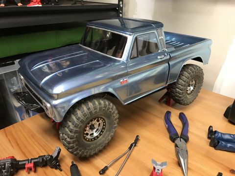 1989 Ford Bronco - A blue RC pickup truck model is elevated on jack stands, surrounded by various tools on a workbench.
