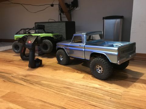 1989 Ford Bronco - Two remote-controlled trucks are parked on a wooden floor, with a controller in the foreground.