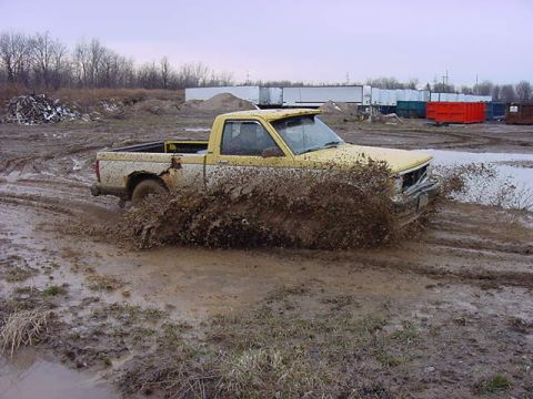 1994 Ford Bronco - A yellow pickup truck navigates a muddy terrain, creating a large spray of mud as it moves.