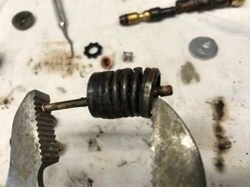 Close-up of a dirty carburetor spring and plunger assembly held by pliers, with other small parts on a white cloth.