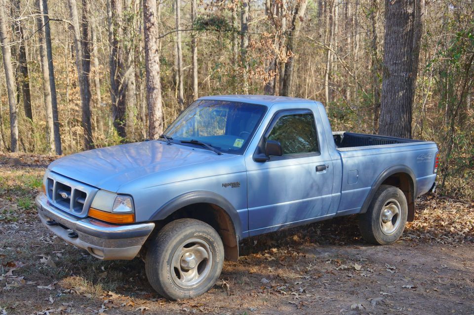 Light blue 1998 Ford Ranger regular cab pickup truck parked on a dirt road in a wooded area.