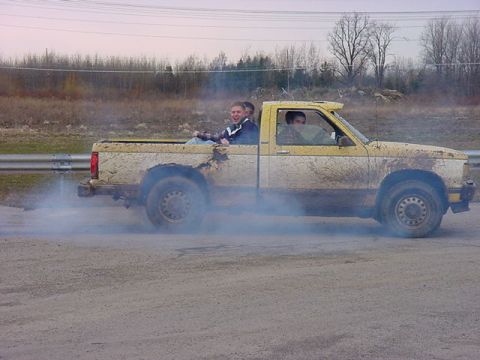 1994 Ford Bronco - A yellow pickup truck, covered in mud, drifts with passengers in the bed.