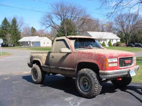 1994 Ford Bronco - This red GMC pickup truck is heavily covered in mud, obscuring much of its original paint and details. The front grille suggests a 1995 mod…