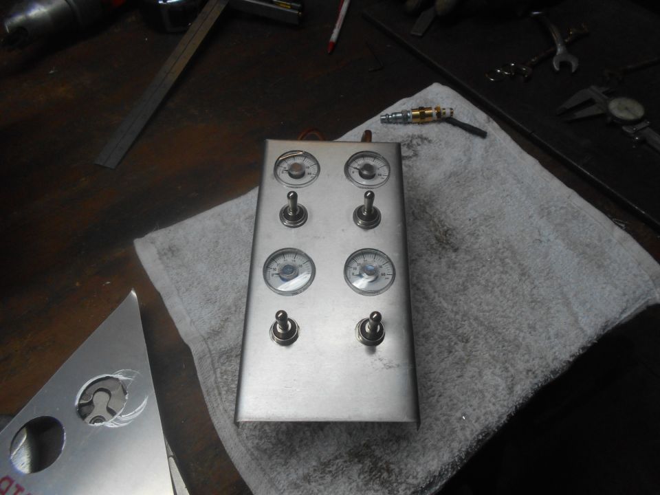 Metal control panel with gauges and toggle switches on a workbench.