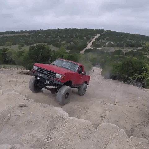 1989 Ford Bronco - A red SUV navigates a dusty, uphill trail surrounded by dry vegetation and overcast skies.